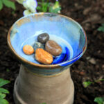 CERAMIC BOWL PLACED ON TOP OF AN UPSIDE DOWN FLOWER POT, CONTAINING WATER AND COLOURFUL RIVER STONES